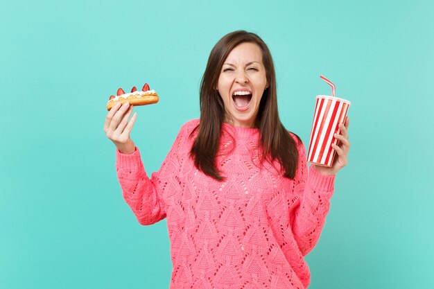 Mujer joven loca en suéter rosa de punto gritando, sostenga en las manos pastel de eclair, vaso de plástico de cola o refresco aislado sobre fondo azul, retrato de estudio. Concepto de estilo de vida de personas. Simulacros de espacio de copia.