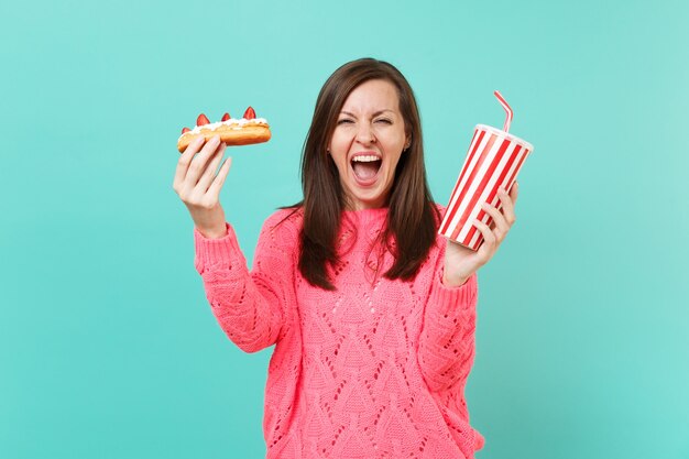 Mujer joven loca en suéter rosa de punto gritando, sostenga en las manos pastel de eclair, vaso de plástico de cola o refresco aislado sobre fondo azul, retrato de estudio. Concepto de estilo de vida de personas. Simulacros de espacio de copia.