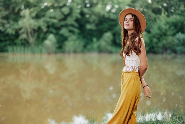 Una mujer joven con un look hippie viaja en la naturaleza junto al lago con un sombrero y pantalones amarillos en otoño