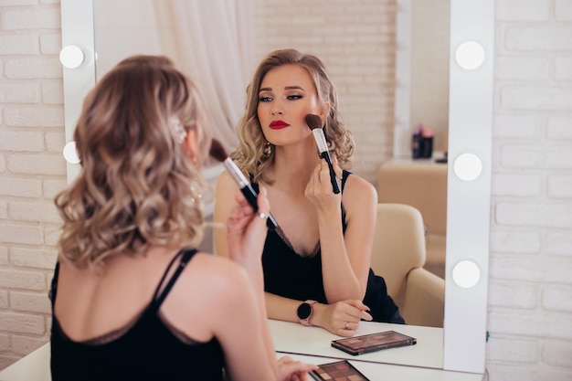 Mujer joven de lujo con peinado rubio y maquillaje mirando al espejo en la peluquería