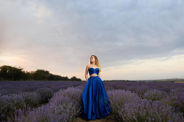 Mujer joven en un lujoso vestido azul de pie en un campo de lavanda contra el fondo del cielo.