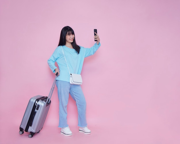 Mujer joven con maleta y smartphone en pared rosa