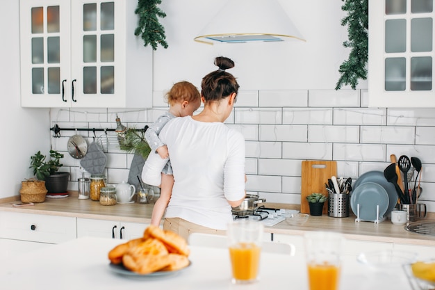 Mujer joven mamá con niña en manos cocinar el desayuno en la cocina brillante en casa