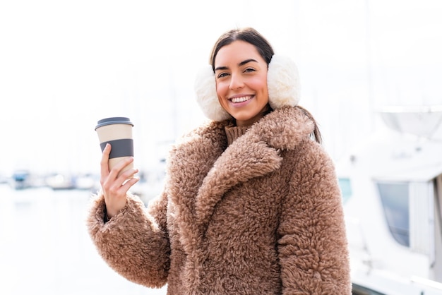 Mujer joven con manguitos de invierno y sosteniendo café para llevar al aire libre sonriendo mucho