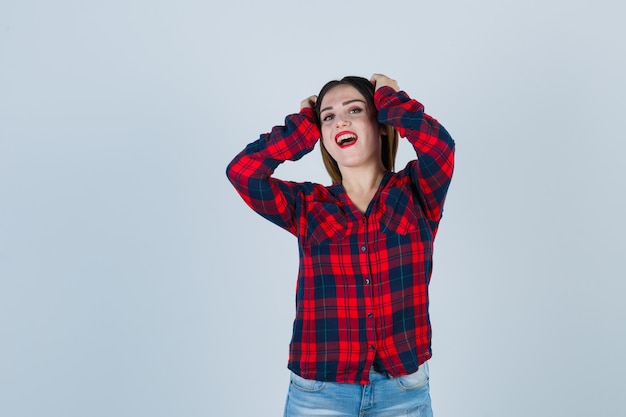 Mujer joven con las manos en la cabeza en camisa a cuadros, jeans y mirando alegre, vista frontal.