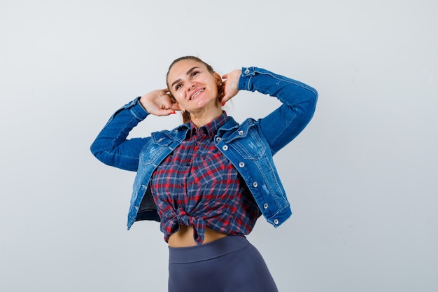 Mujer joven con las manos en la cabeza mientras mira hacia arriba con camisa a cuadros, chaqueta, pantalones y mirando alegre, vista frontal.