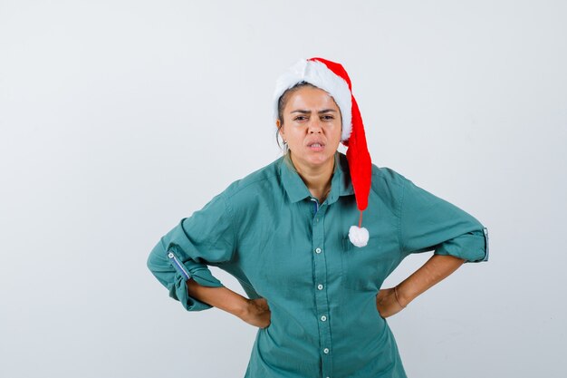 Mujer joven con las manos en la cintura en camisa, gorro de Papá Noel y mirando enojado, vista frontal.