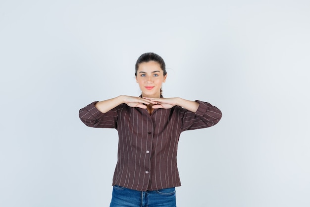 Mujer joven con las manos debajo de la barbilla en camisa a rayas, jeans y aspecto atractivo, vista frontal.