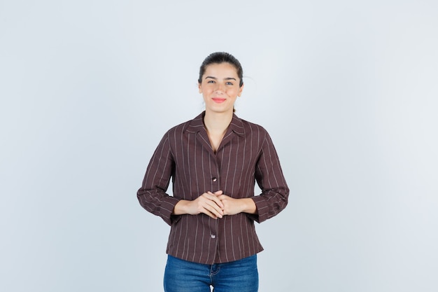 Mujer joven con las manos delante de ella en camisa a rayas, jeans y mirando feliz, vista frontal.