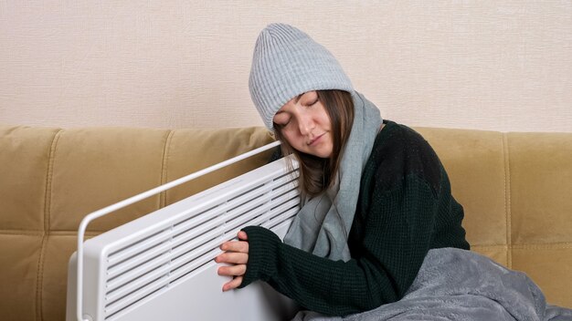 Mujer joven con manta gris en bufanda y gorro de punto abrazos calentador sentado en un sofá moderno y suave en la sala de estar en la fría temporada de invierno