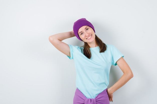 Mujer joven manteniendo la mano detrás de la cabeza en camiseta, gorro y mirando feliz, vista frontal.