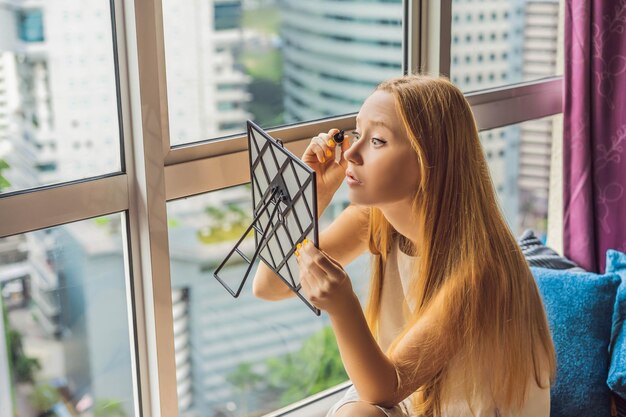 La mujer joven se maquilla sentada junto a la ventana con una vista panorámica de los rascacielos y los grandes