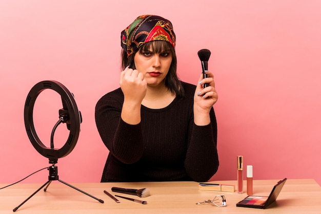 Mujer joven maquilladora haciendo un tutorial de maquillaje aislado sobre fondo rosa mostrando el puño a la cámara expresión facial agresiva