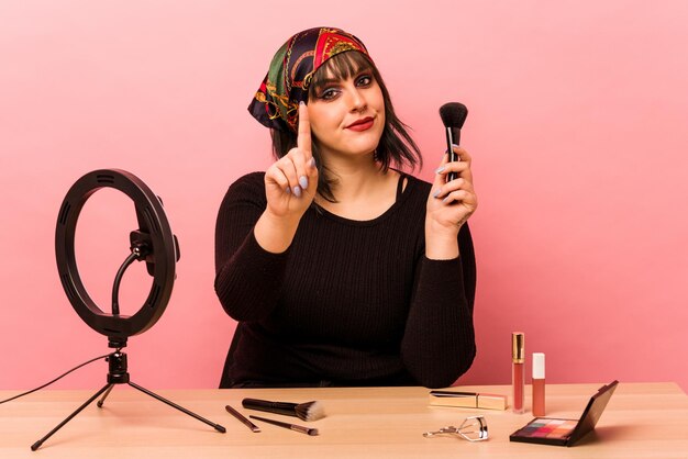 Mujer joven maquilladora haciendo un tutorial de maquillaje aislado sobre fondo rosa que muestra el número uno con el dedo