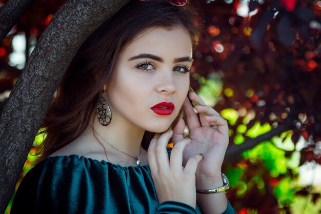 Mujer joven con maquillaje brillante, posando cerca del árbol con hojas rojas.