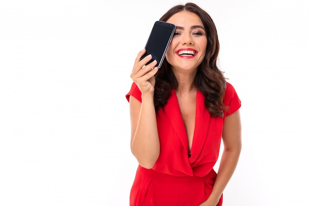 Una mujer joven con maquillaje brillante, con un vestido rojo de verano se para con un teléfono en la mano y sonríe