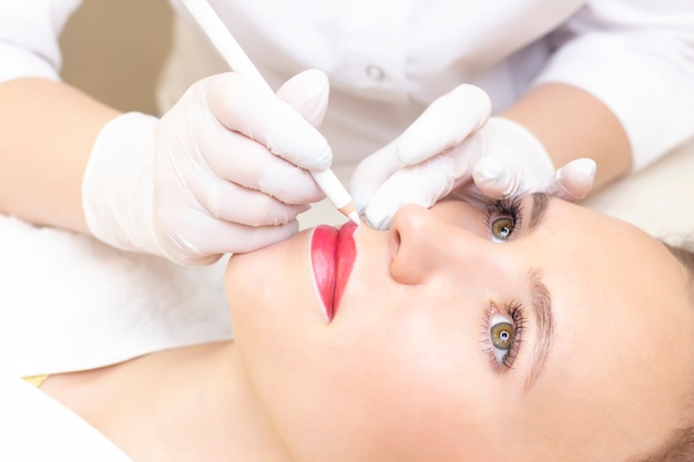 Mujer joven con maquillaje permanente en sus labios en el salón de belleza. Maquillaje permanente (tatuaje). dibujar un contorno con un lápiz labial blanco