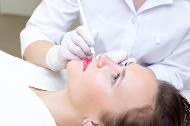 Mujer joven con maquillaje permanente en sus labios en el salón de esteticistas. Maquillaje permanente (tatuaje). dibujar un contorno con un lápiz labial blanco