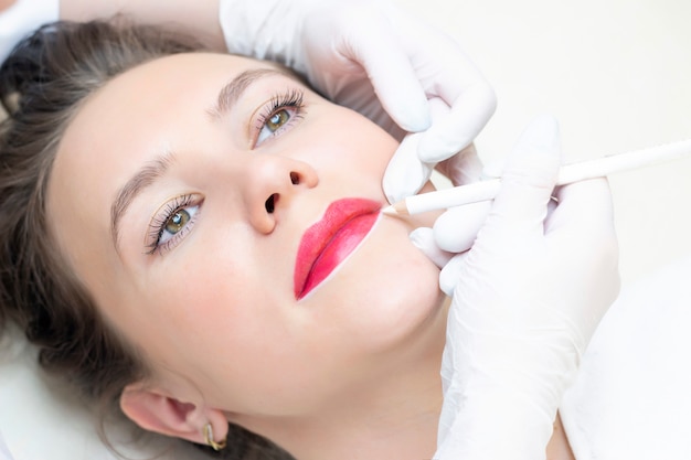 Mujer joven con maquillaje permanente en sus labios en el salón de esteticistas. Maquillaje permanente (tatuaje). dibujar un contorno con un lápiz labial blanco