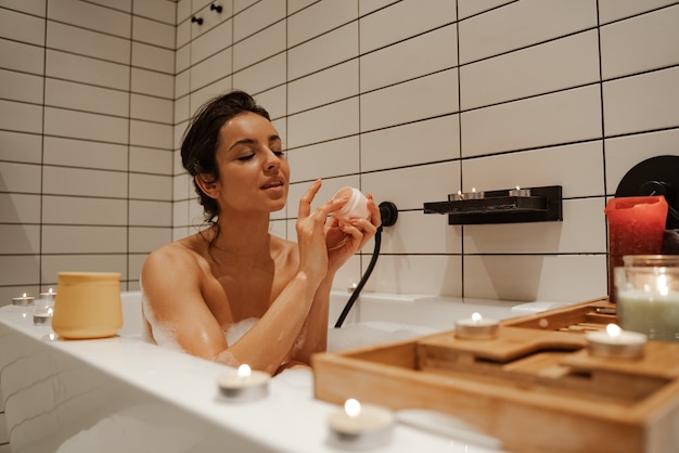 Mujer joven con máscara en la cara, relajándose en el acogedor baño lleno de espuma y decorado con velas. Trato saludable y disfrute.