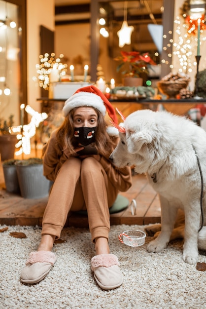 Mujer joven en máscara facial celebrando con un perro vacaciones de año nuevo en casa. Concepto de cuarentena y autoaislamiento durante la epidemia en vacaciones