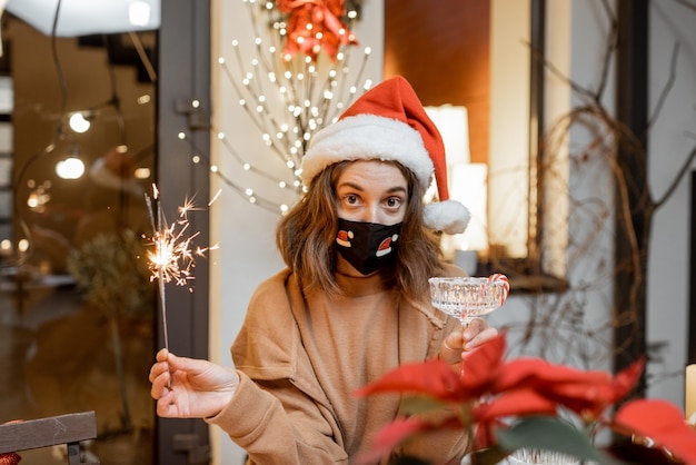 Mujer joven en máscara facial celebrando solo las vacaciones de año nuevo en casa. Concepto de cuarentena y autoaislamiento durante la epidemia en vacaciones