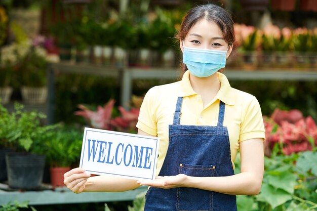 Mujer joven con máscara médica al invitar a los clientes al centro de jardinería o al vivero de plantas