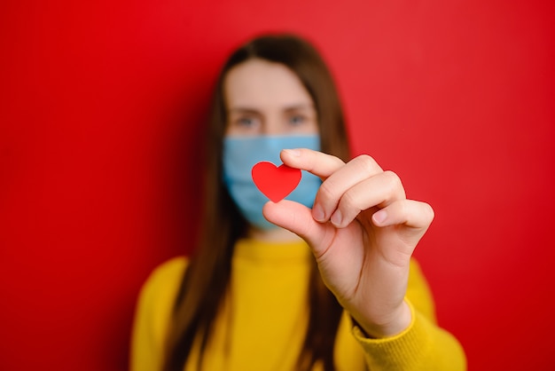 Mujer joven con una máscara médica azul protectora, aislada sobre fondo rojo, sosteniendo el corazón rojo como una forma de mostrar agradecimiento y agradecer a todos los empleados esenciales durante las pandemias de Covid-19. Enfoque selectivo