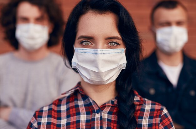 Mujer joven con máscara médica mirando a cámara sobre fondo borroso de amigos durante la epidemia de coronavirus