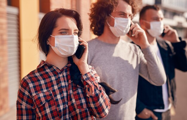 Foto mujer joven con máscara médica mirando a otro lado y teniendo una conversación por teléfono inteligente mientras está de pie en la calle de la ciudad cerca de los hombres durante la pandemia de coronavirus