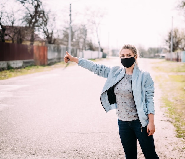 Foto una mujer joven con una máscara médica negra se encuentra en la carretera con una mano levantada