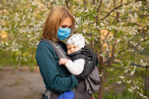 Mujer joven en máscara médica con un niño en sus brazos Alegría de la maternidad y el desarrollo infantil árbol de flores a fines de la primavera