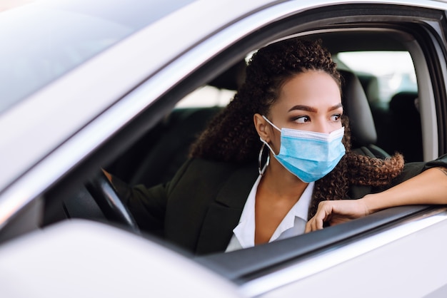 Mujer joven con máscara médica protectora conduciendo un coche.