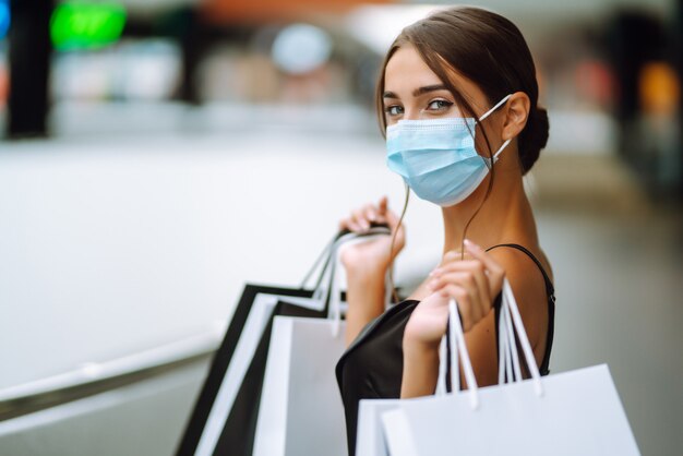 Foto mujer joven en máscara médica protectora estéril en su rostro con bolsas de compras en el centro comercial.