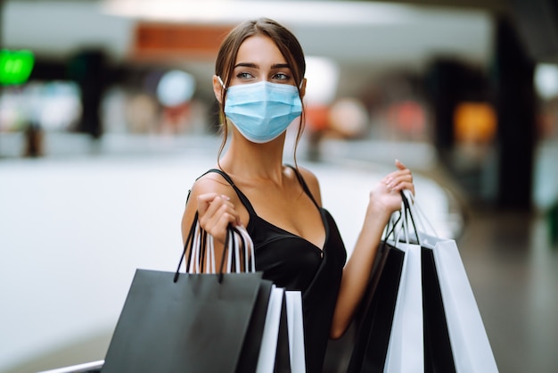 Mujer joven en máscara médica protectora estéril en su rostro con bolsas de compras en el centro comercial.