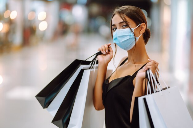 Mujer joven en máscara médica protectora estéril en su rostro con bolsas de compras en el centro comercial.