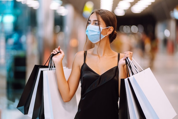 Mujer joven en máscara médica protectora estéril en su rostro con bolsas de compras en el centro comercial.