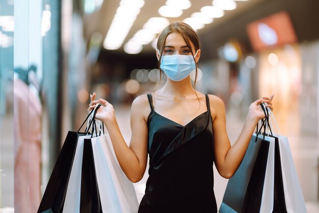 Mujer joven en máscara médica protectora estéril en su rostro con bolsas de compras en el centro comercial.