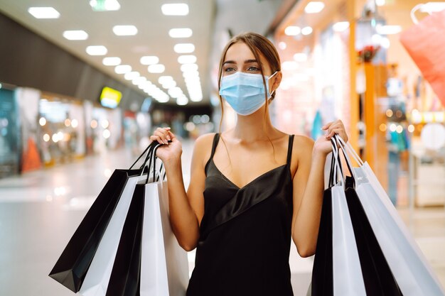 Mujer joven en máscara médica protectora estéril en su rostro con bolsas de compras en el centro comercial.