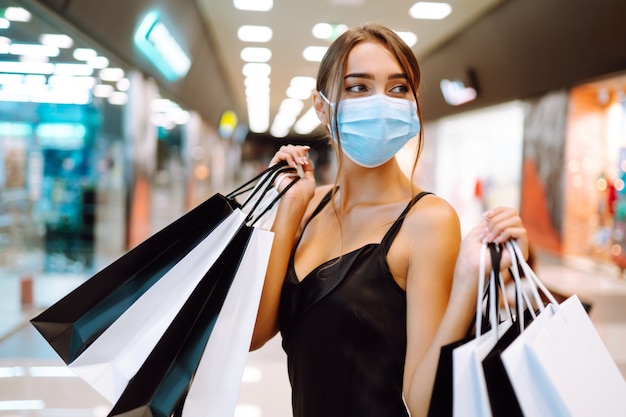 Mujer joven en máscara médica protectora estéril en su rostro con bolsas de compras en el centro comercial.