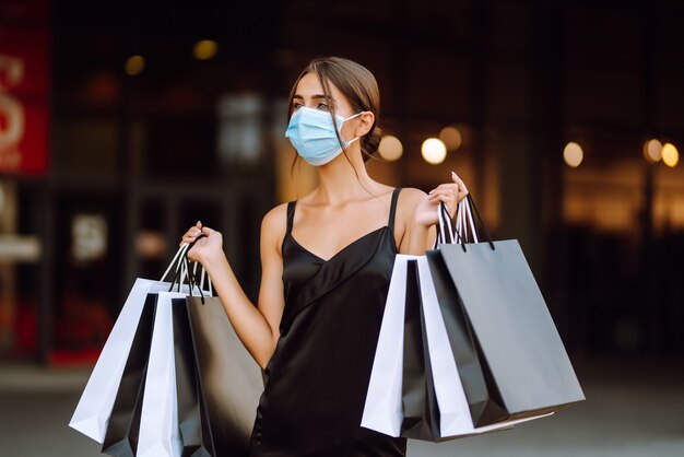 Mujer joven en máscara médica protectora estéril en su rostro con bolsas de compras disfrutando de compras.