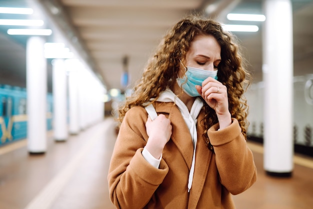 Mujer joven en máscara médica protectora estéril tosiendo en la estación de metro.