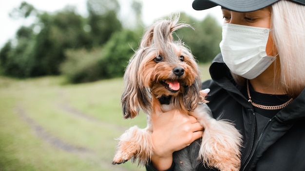 Mujer joven con una máscara médica tiene un perro pequeño en sus brazos