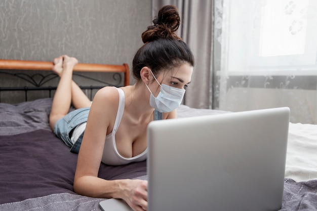 Una mujer joven con una máscara médica trabaja desde su casa en una computadora portátil.