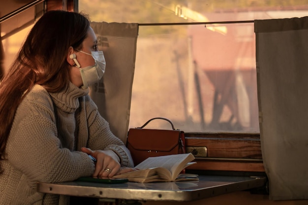 Una mujer joven con una máscara médica viaja sola en tren Tomando precauciones en el transporte público La niña está leyendo un libro y escuchando música en el tren