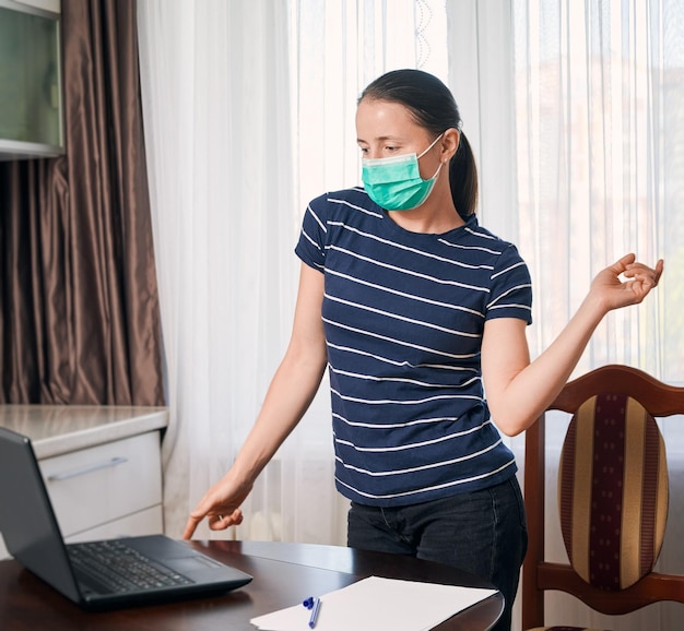 Mujer joven con máscara de pie en su lugar de trabajo en casa frente a su computadora portátil haciendo ejercicios para calentarse después de un largo día de trabajo Auto-aislación durante el tiempo de crisis Quédese en casa concepto de salvar vidas