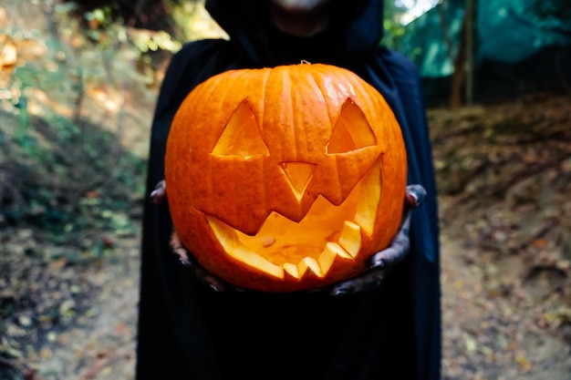 Mujer joven con máscara de pintura de halloween con capucha negra Bruja aterradora sosteniendo calabaza ansiada espeluznante