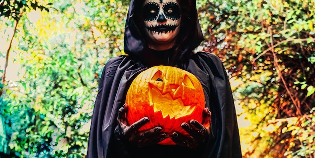 Mujer joven con máscara de pintura de halloween con capucha negra Bruja aterradora sosteniendo calabaza ansiada espeluznante