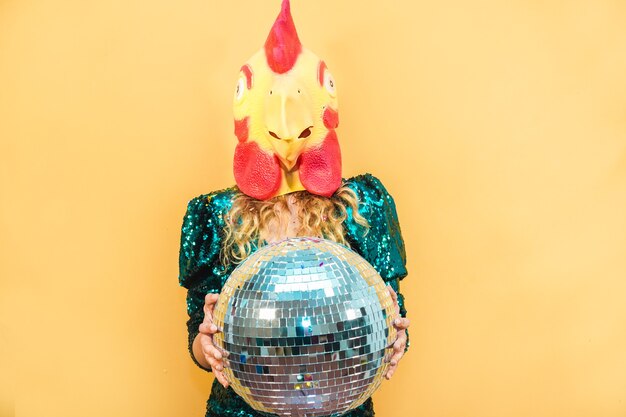 Mujer joven con máscara de pollo mientras sostiene la bola de discoteca en la fiesta de año nuevo - Enfoque suave en la cara