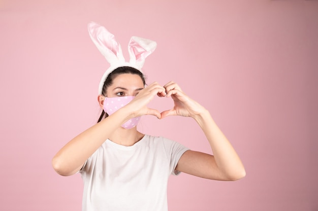 Mujer joven con máscara de protección contra el coronavirus para la epidemia de covid-19 haciendo forma de símbolo de corazón con las manos en rosa